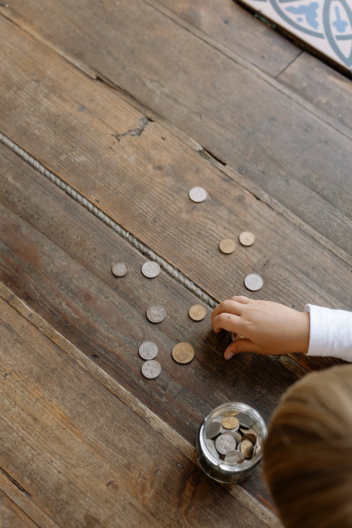 Overhead view of childs hand gathering coins into a glass jar on wooden flooring.